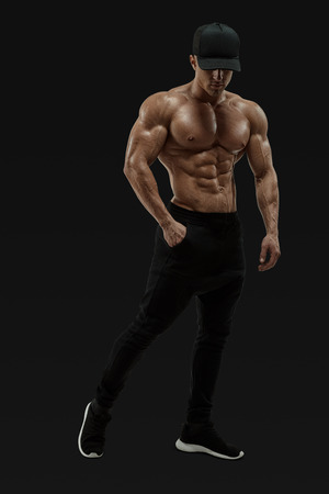 Full-length portrait of shirtless male bodybuilder with muscular physique posing with muscular build strong abs showing. Masculine young fitness male model posing shirtless against black background. Vector mask clipping pathの写真素材