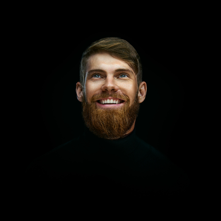 Close-up portrait of laughing young bearded man weared black turtleneck looking up Studio shot over black background.の写真素材