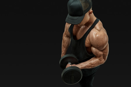 Close up shot of muscular bodybuilder wearing a red tank top and black cap doing biceps curl with dumbbell against black background. Image with clipping pathの写真素材