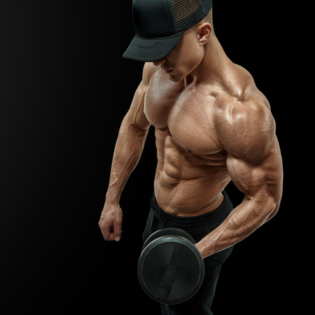 Closeup of a handsome power athletic man bodybuilder doing exercises with dumbbell in left hand. Strong bodybuilder with six pack, perfect abs, shoulders, biceps, triceps and chest. Wearing a black baseball cap. Vector path clipping maskの写真素材
