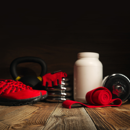 Blurred image Sport fitness bodybuilding healthy lifestyle and weightlifting concept gym background Dumbbell plates White jar with whey protein Red hand wrap and red gloves on wooden floorの写真素材