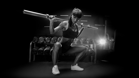Black and white image of fit young woman in great shape lifting barbells looking down, working out in a gym Sporty sexy woman doing squat workout Strong legsの写真素材