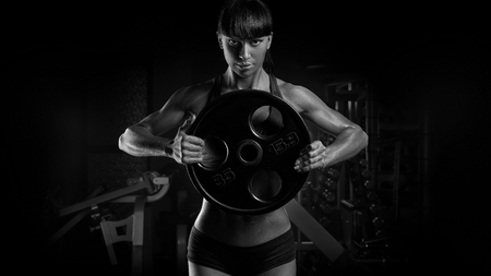 Black and white photo of fit power athletic confident young woman bodybuilder doing exercises with heavy weight barbell plate in gym. Fitness muscular body, strong hand on dark background Pumping up musclesの写真素材
