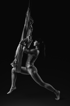 Black and white image of young woman posing while doing anti-gravity aerial yoga in gray hammock isolated on black background Clipping pathの写真素材