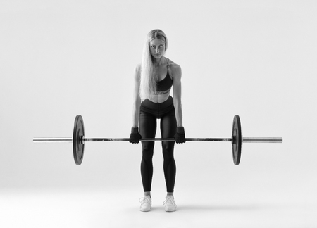 Black and white Front view of confident young blonde woman doing weight lifting workout Attractive young woman bodybuilder lifting barbells looking forward Strong trained body shape arms chest legsの写真素材