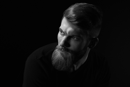 Dramatic concept Black and white close-up portrait of young handsome bearded man looking forward. Studio shot on black backgroundの写真素材