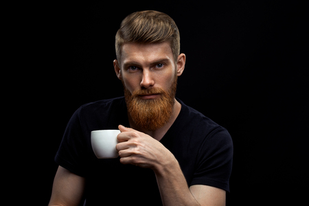 Beautiful young bearded man drinking coffee and looking at camera Resting handsome perfect hairstyle man drinking espresso coffee holding cup of coffee in hand Studio shot on black background.の写真素材