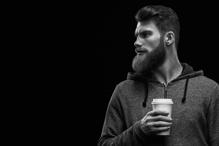 Black and white dramatic light studio shot of young caucasian bearded stylish man with cup of coffee Handsome hairstyling male portrait holding coffee to go cup over black background .の写真素材
