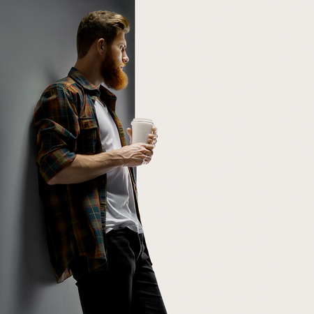 Portrait of handsome single bearded young man with serious expression having coffee indoors and white short t-shirt looking over white background with free copy spaceの写真素材