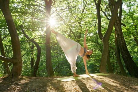 Young yoga asian attractive woman practicing yoga concept, standing in Half Moon exercise, Ardha Chandrasana pose, working out, wearing sportswear Practicing yoga in the nature at sunsetの写真素材