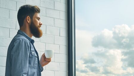 Portrait of a bearded young man drinking morning coffee. A bearded man with a mustache looks out the large window.の写真素材
