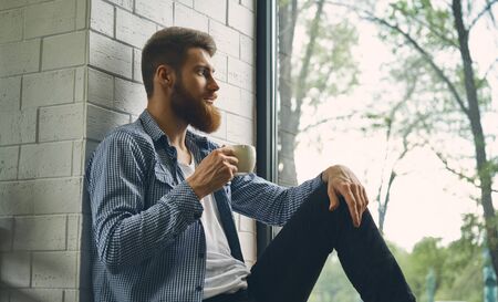 Young attractive bearded man rests and looks out the window. Enjoy the morning afternoon. Healthy lifestyle concept. Drink fresh bracing coffee looking into the distanceの写真素材