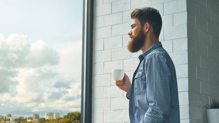 Attractive bearded young man is enjoying hot coffee. Portrait of handsome bearded caucasian man with cup at modern coffee shop. Guy in cafe relaxing. Freelance people lifestyle with copy space conceptの写真素材