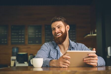 Elegant hipster freelancer multitasking multimedia man using devices Remote job concept Happy handsome bearded man using a digital tablet at cofe smiling laughsの写真素材