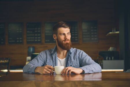 Young attractive man holding coffee cup drinking morning espresso coffee Bearded man is tasting coffee while restingの写真素材