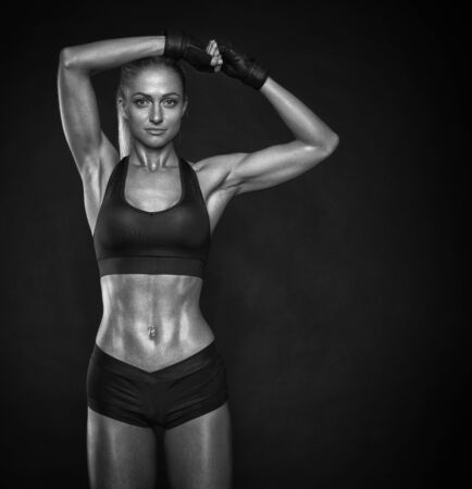 Black and white image of fitness woman in sports bra. She is looking at the camera and holds her hands above her head. Beautiful woman with clean and glowing skin isolated on dark backgroundの写真素材