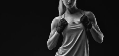 Black and white horizontal portrait of young woman with muscular body standing against black background. Image of fitness woman in sports clothing and gloves.の写真素材