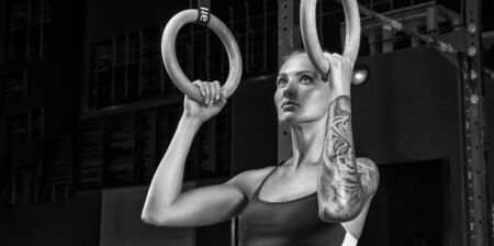 Fitness girl exercising with gymnastic rings in gym. Attractive Female athlete standing at gym holding dip rings and looking up. Woman training olympic ring in crossfit room. Black and white photo.の写真素材