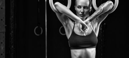 Strong young woman in a black sports top holding gymnast rings at the gym and looking down Young female exercise with dip rings. Healthy lifestyle, sport black and white wide close-up cover conceptの写真素材