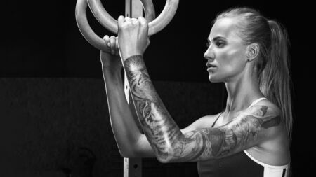 Female fitness athlete in black sportswear holding dip rings and looking away Girl exercising with gymnastic rings in gym. Caucasian woman training olympic ring in crossfit room Black and white photo.の写真素材