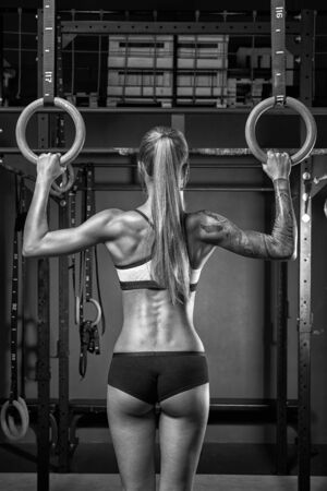 Back view of fit young caucasian woman with ponytail in sports wear Healthy woman exercising with gymnastic rings Female bodybuilder with power hand, triceps, arms, rear in gym. Black and white photo.の写真素材