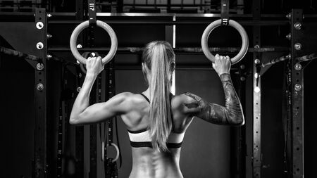 Back view of fit young woman with ponytail in sports wear Healthy woman exercising with gymnastic rings Female bodybuilder with power hand, triceps, arms, rear in gym Black and white horizontal photo.の写真素材