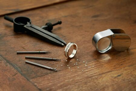 Jeweler workplace with golden ring, tools and equipment for working on an old wooden table. Ready-made diamond engagement ring, diamonds, magnifying glass. Engraver at work on jewelry from diamondsの写真素材