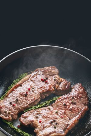 Steaming, reeky, flavored spicy grilled beef striploin steak in a frying pan on a black background with copy space. Frying pan, steak, cooking on fire conceptの写真素材