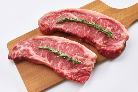 Raw juicy beef steak ready to be cooked. Close up photo of two fresh raw striploin steak on wooden board on white background with rosmary. Top view.の写真素材
