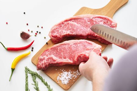 Man hands slicing raw beef steak with knife on wood cutting desk surrounded by ingredients for preparing the meat. Top view food preparation process concept.の写真素材