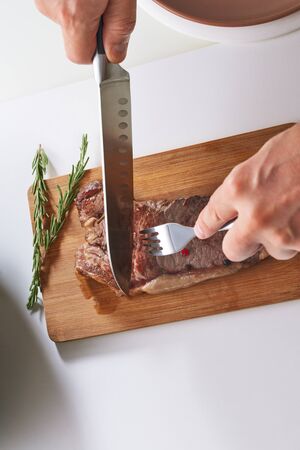 Man cutting grilled steak on wooden board in kitchen. Close up sliced medium rare grilled beef steak with salt, pepper and rosemary on meat cutting board on white backgroundの写真素材