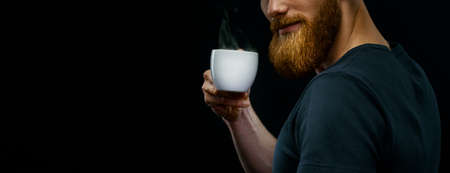 Cup of hot espresso coffee in the hand of a young bearded guy. Close-up studio shot on black background. Copy free space on left.の写真素材