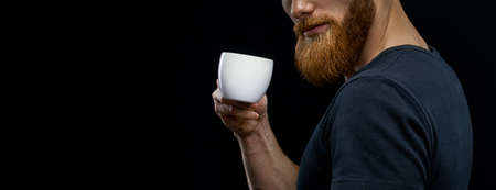 Closeup shot of coffee cup. Beautiful young bearded man drinking coffee. Resting bearded man drinking espresso coffee holding cup of coffee in hand Studio shot on black backgroundの写真素材