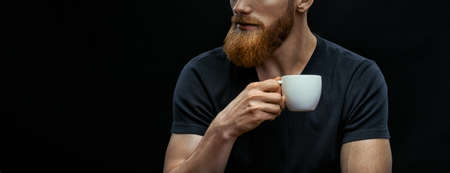 Croped shot of bearded man drinking coffee. Resting man drinking espresso coffee holding cup of coffee in hand. Studio shot on black background wit copy space on leftの写真素材