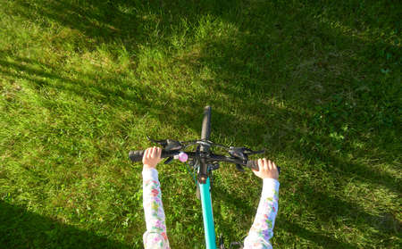 Focus on grass. Top view of hands of a girl holding the bicycle handlebar of a blue bicycle. Teenage girl rides a bicycle in the park on bright green grass on a sunny summer day.の写真素材