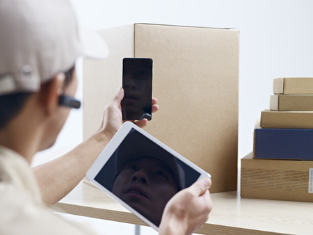 male asian courier company worker using cellphone and tablet computer to scan information on packages.の写真素材