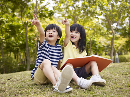 little asian boy and girl with book in hand sitting on grass pointing to the sky.の写真素材