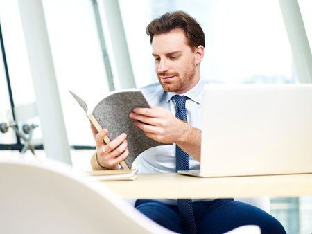 caucasian business man looking through notes while working on laptop computer in office.の写真素材