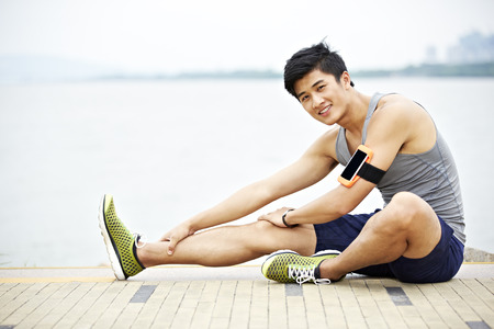 young handsome asian jogger wearing fitness tracker stretching a leg by lake looking at camera smilingの写真素材