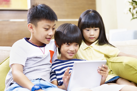 three asian children sitting on couch at home playing video game using digital tablet.の写真素材