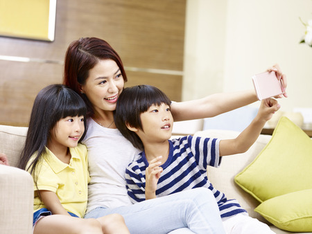 young asian mother and children sitting on sofa taking a selfie with mobile phone, happy and smiling.の写真素材