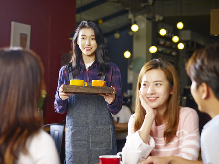 young smiling asian waitress serving customers in coffee shop, focus on the background girl.の写真素材