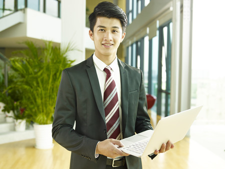 ortrait of a young asian business man, holding laptop computer, looking at camera smilingの写真素材