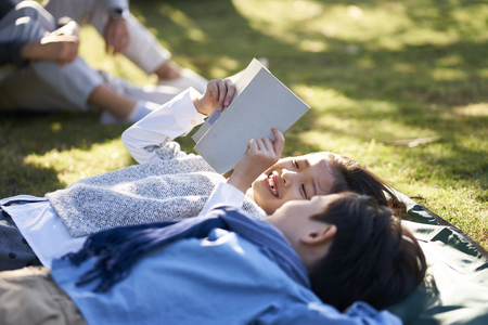 two little asian kids boy and girl lying on back on grass in park reading a book together with parents sitting in background.の写真素材