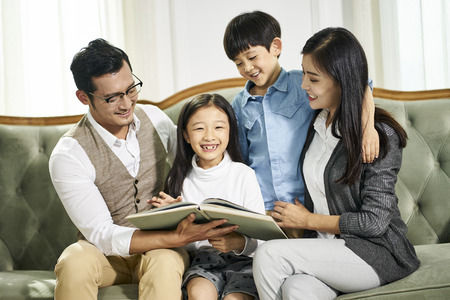 Young asian parents and two children sitting on couch reading book together in living room at homeの写真素材