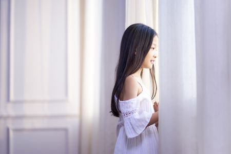 beautiful little asian girl with long black hair looking out of window in bedroomの写真素材