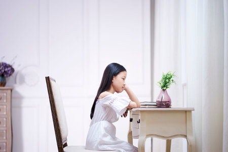 sad little asian girl with long black hair sitting at desk in her room thinking with hand on chinの写真素材