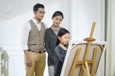 Beautiful little asian girl with long black hair drawing a painting on canvas while parents standing behind watching.の写真素材