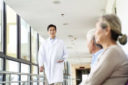 young asian doctor walking in hospital hallway to meet patients in waiting areaの写真素材