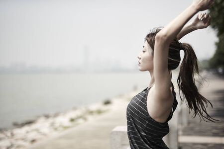 young asian adult woman stretching arms outdoors by the sea, side viewの写真素材